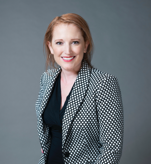 Portrait of Meghan Race smiling, wearing a black-and-white patterned blazer over a black top, photographed against a neutral gray studio background.